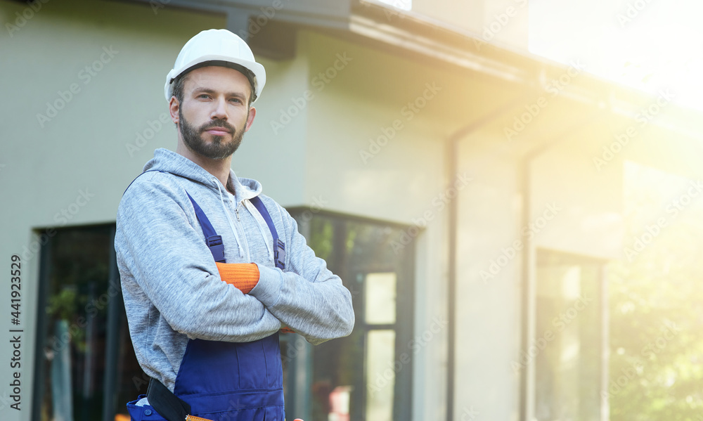 Portrait of young bearded male engineer in hard hat looking at camera, posing outdoors while working on cottage construction