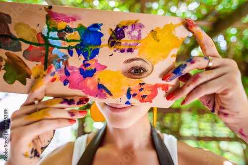Fototapeta Naklejka Na Ścianę i Meble -  Playful portrait of a young gorgeous female artist painter covered in paint, looking and smiling at camera through her painter's palette. Creativity and individuality concept.