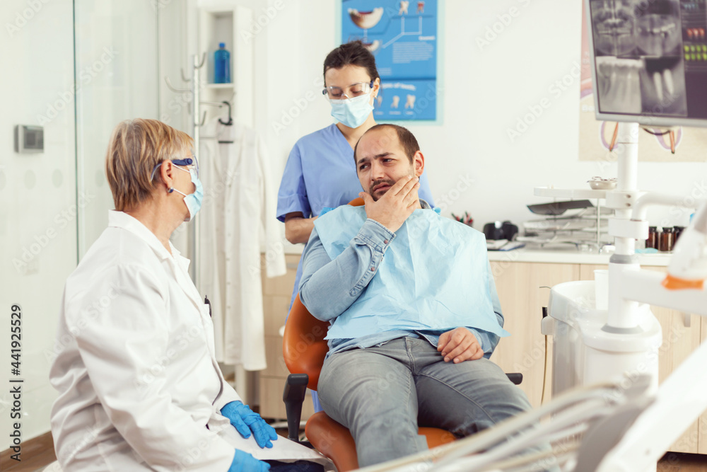 Obraz premium Sick patient complaining about toothache while talking with dentist before intervetion. Senior doctor speaking to man sitting on stomatological chair while nurse stomatology hospital office