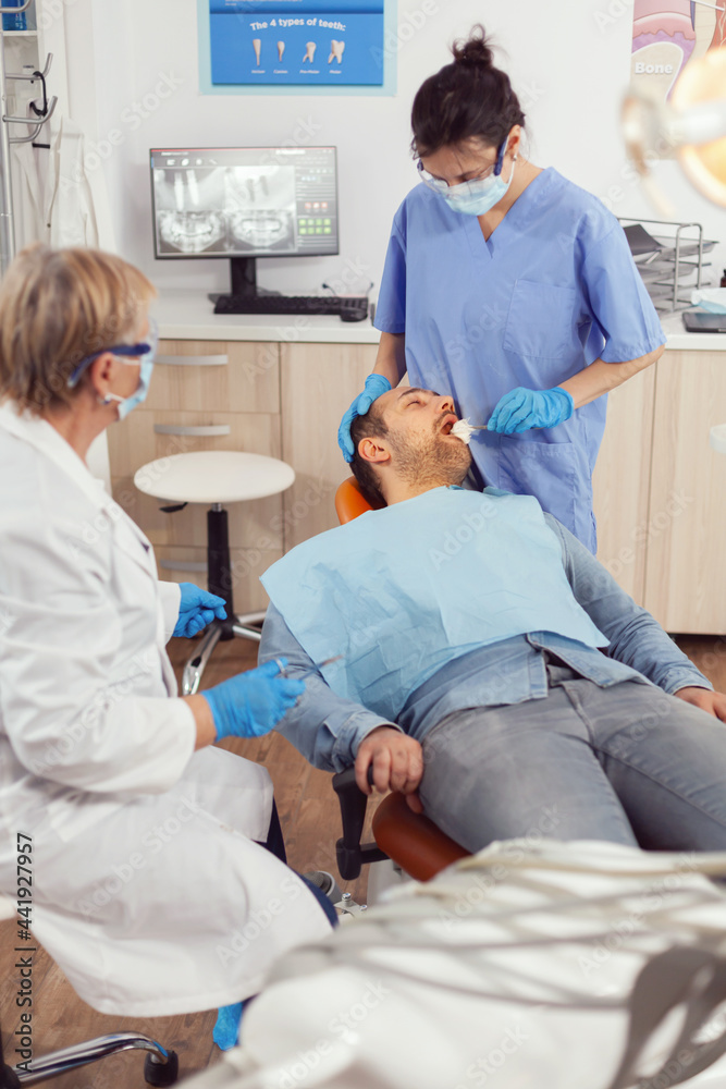 Senior stomatologist woman preparing for surgery putting implant to sick patient mouth in stomatological hospital. Dentist doctor with protection mask treating teeth of man sitting on dental chair Senior stomatologist woman preparing for surgery putting implant to sick patient mouth in stomatological hospital. Dentist doctor with protection mask treating teeth of man sitting on dental chair