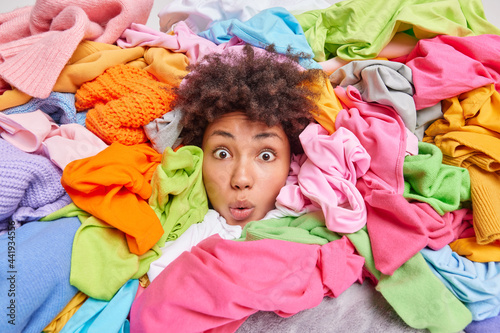 Decluttering second hand spring cleaning fast fashion and organization of life. Stunned Afro American woman with curly hair looks through big heap of colorful clothes puts things in order at closet photo
