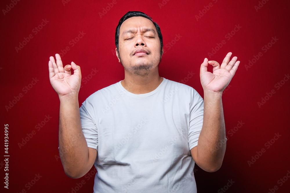Young asian man wearing white t-shirt relax and smiling with eyes closed doing meditation gesture with fingers. Yoga concept. Young asian man wearing white t-shirt relax and smiling with eyes closed doing meditation gesture with fingers. Yoga concept.