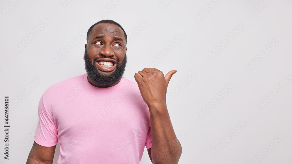 That way. Positive dark skinned man with white teeth points thumb away on blank space shows place for logo or product placement dressed in casual pink t shirt isolated over white background. That way. Positive dark skinned man with white teeth points thumb away on blank space shows place for logo or product placement dressed in casual pink t shirt isolated over white background.