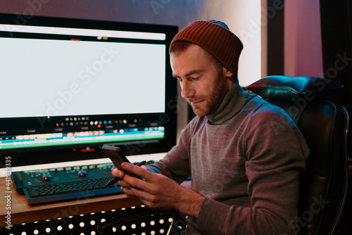 Attractive Male Video Editor Works with Footage or Video on His Personal Computer and having a break communicating on his smartphone. He Works in Creative Office Studio or home. Neon lights photo