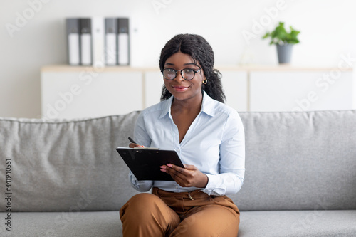 Portrait of positive black female psychologist looking at camera and taking notes during consultation at office