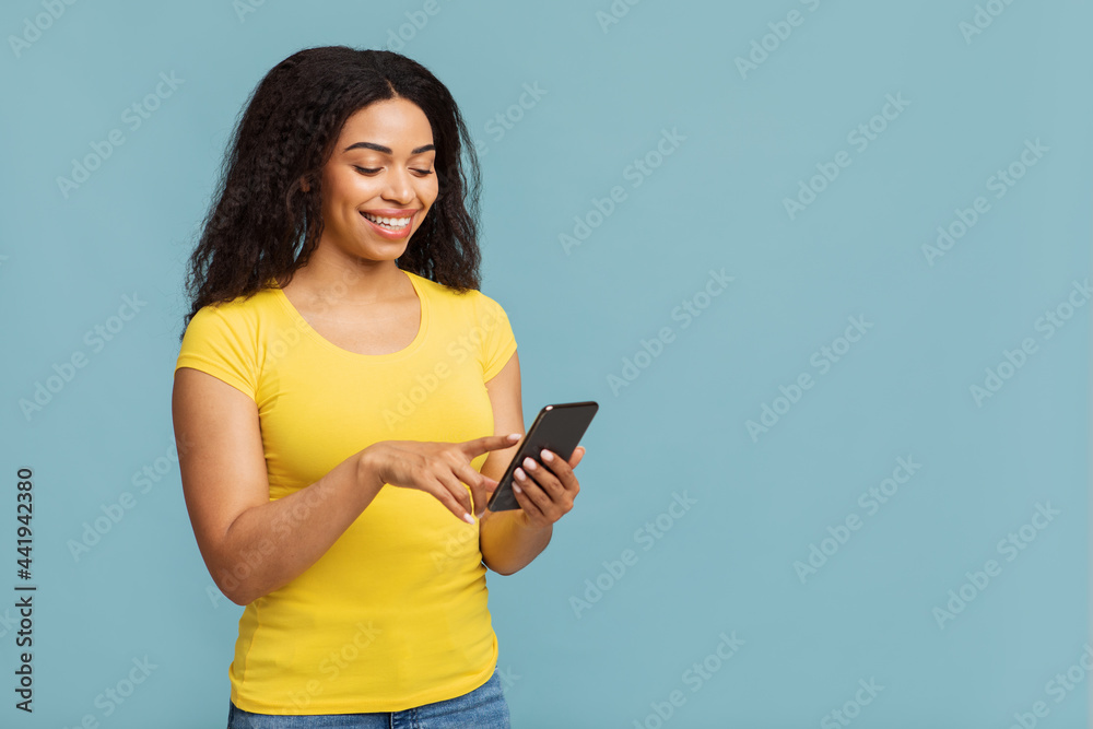 Cool app. Happy african american woman using smartphone, standing over blue studio background with free space Cool app. Happy african american woman using smartphone, standing over blue studio background with free space
