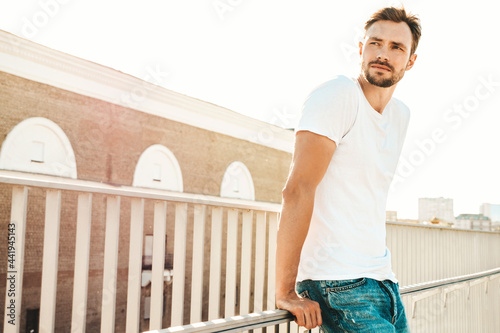 Portrait of handsome smiling hipster lambersexual model.Stylish man dressed in white T-shirt and jeans. Fashion male posing on the street background near fence at the beach at sunset