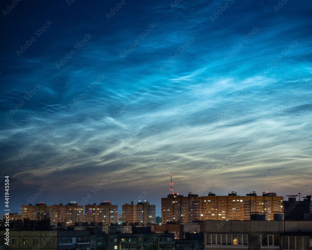 Atmospheric phenomenon glowing of noctilucent silvery clouds in the night shining sky. Silhouettes of city buildings against a background of beautiful luminous clouds.