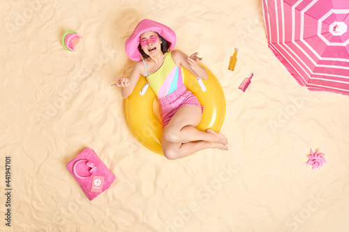 Overhead shot of depressed crying woman feels very upset dressed in summer clothes protective shades poses at sandy beach on inflated swimring during hot day. People resort sunbathing concept
