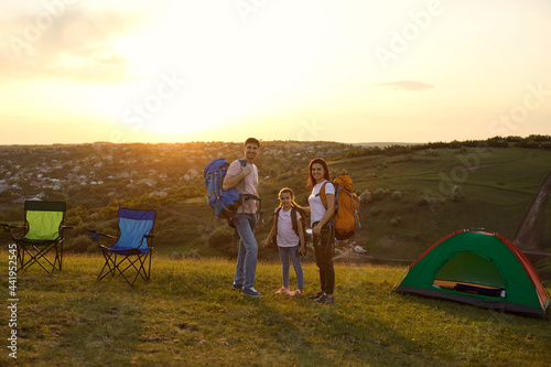 Happy family on summer camping trip at mountains. Adorble little girl and parents with backpacks standing at campground during sunset