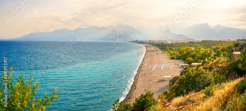 Beautiful wide panorama of beach in Turkish resort Antalya. White line surf separates calm turquoise sea and Konyaalti beach with green parks and chain mountains in rays of evening sun at sunset . photo
