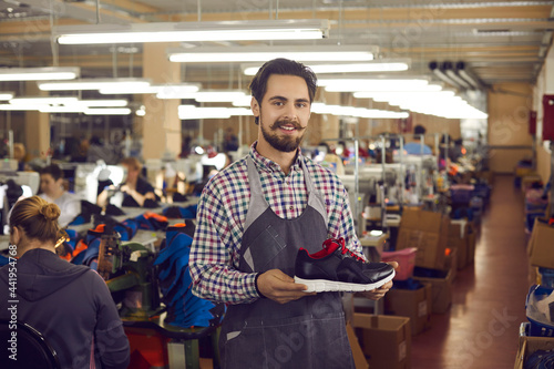 Young male shoemaker designerwith friendly smile on face wearing apron presenting new sports sneakers shoe collection. Trendy modern footwear production, logistics and distribution photo