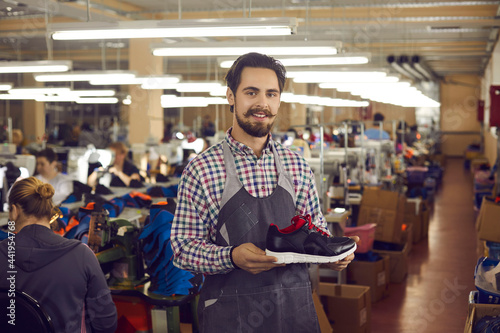 Young male shoemaker designerwith friendly smile on face wearing apron presenting new sports sneakers shoe collection. Trendy modern footwear production, logistics and distribution
