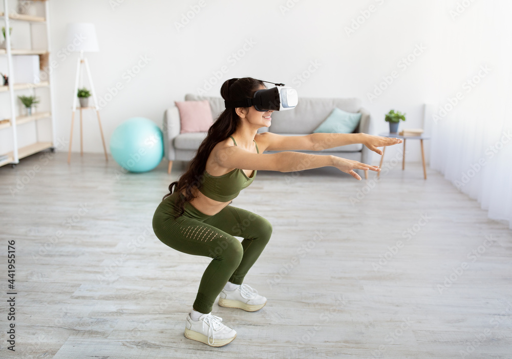 Fototapeta premium Home workout with virtual reality. Young Indian woman in VR headset doing squats at home