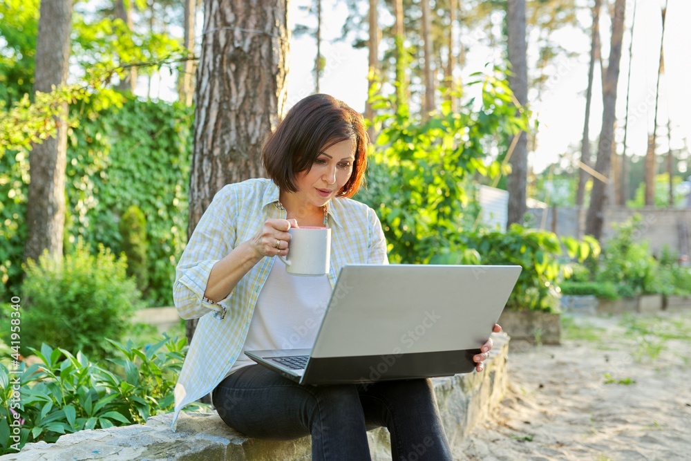 Beautiful emotional middle aged woman relaxing in garden with cup of tea and laptop Beautiful emotional middle aged woman relaxing in garden with cup of tea and laptop