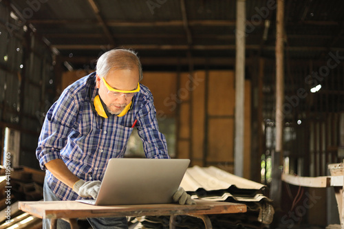 Senior Asian carpenter man is using computer laptop inside his home garage to get new modern inspiration and learning skill from online class and contacting international customers