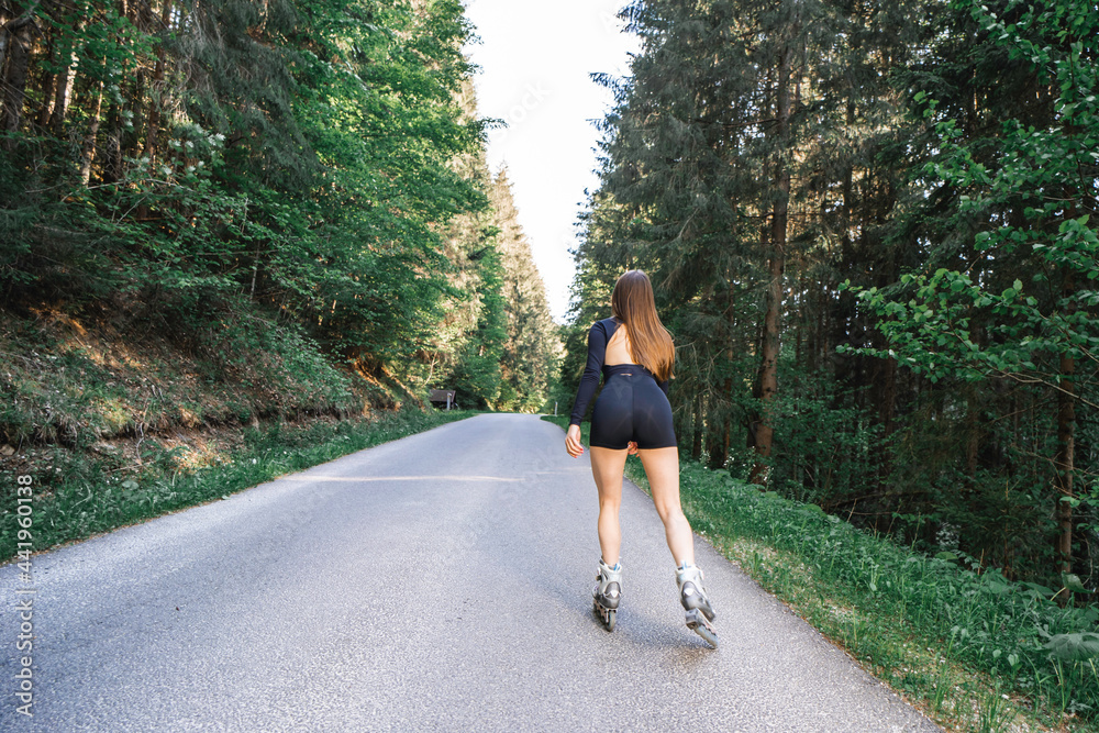Fototapeta premium Young caucasian fit woman on roller skates riding outdoors in woods on asphalt road. Wearing open crop top and black sporty shorts. Shot from behind.