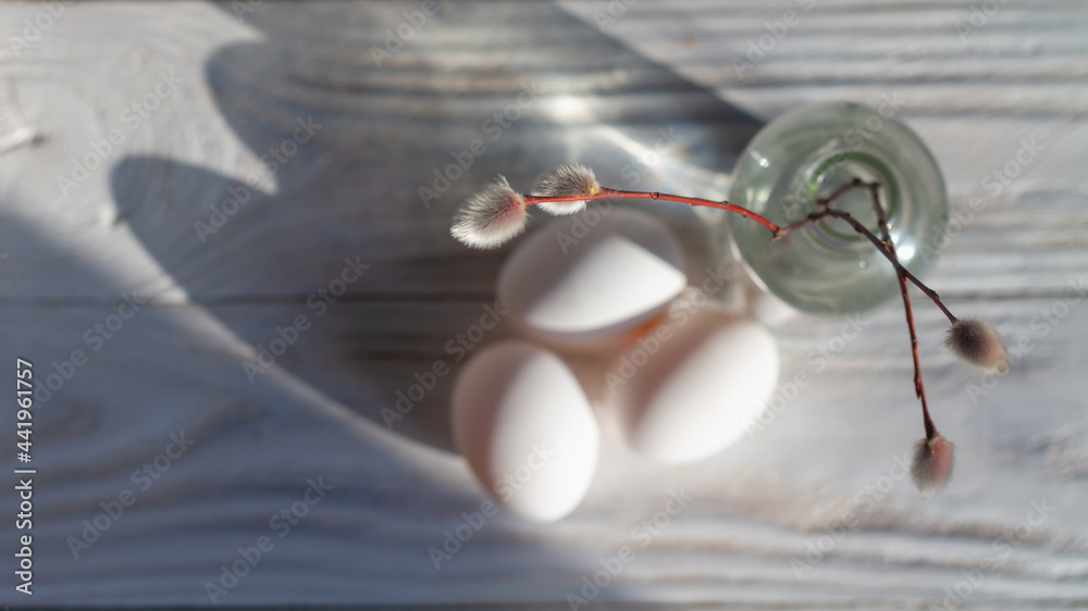 A sprig of willow with fluffy silver buds in a glass bottle and white eggs on the white table at home in the spring for Easter. Top view. Copy space A sprig of willow with fluffy silver buds in a glass bottle and white eggs on the white table at home in the spring for Easter. Top view. Copy space