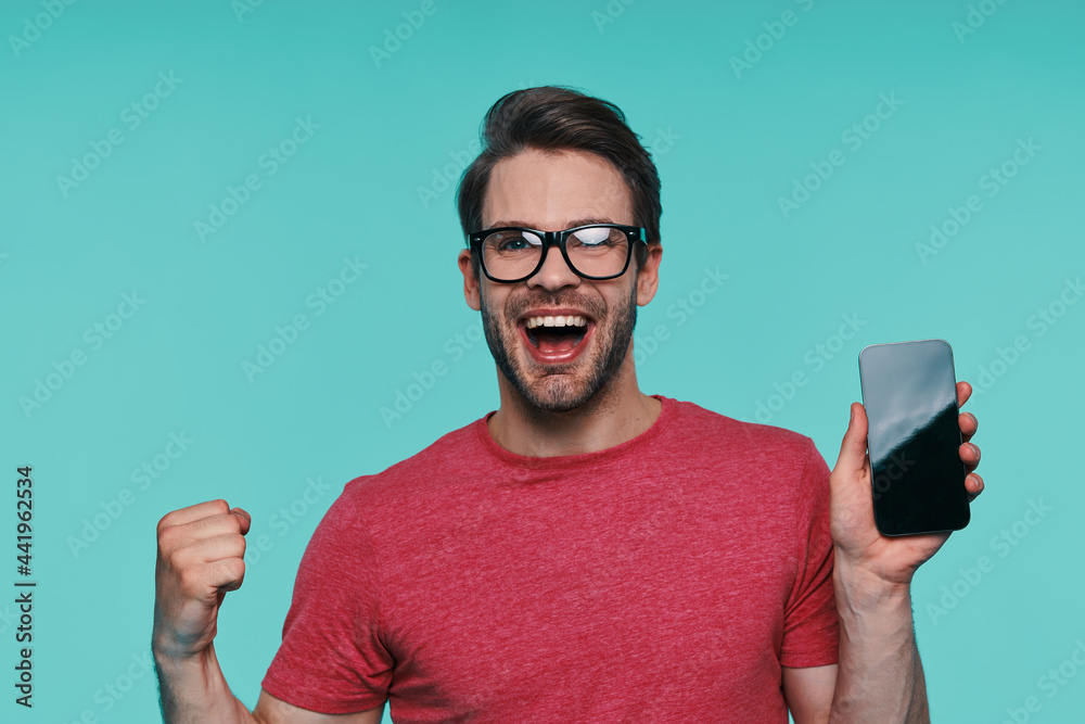 Portrait of handsome young smiling man in eyeglasses showing his smart phone and gesturing Portrait of handsome young smiling man in eyeglasses showing his smart phone and gesturing