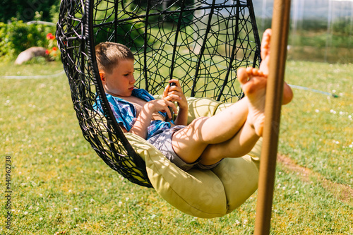 Boy in plaid shirt sitting in cocoon chair and playing with telephone. Smartphone games. Summer time photo