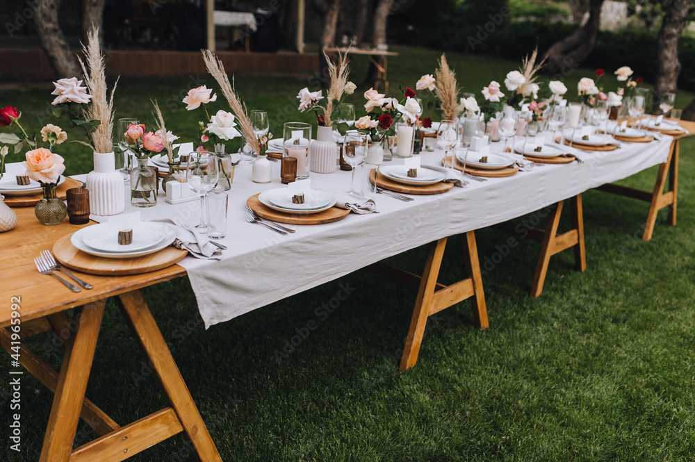 Wooden, long decorated with flowers, reeds, empty dishes, plates, forks, knives, glasses with a white tablecloth stands on the green grass in the park, forest. Wedding banquet.