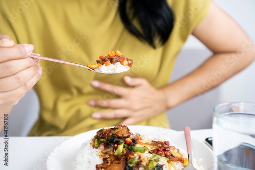 Asian woman eating spicy food and having acid reflux or heartburn hand holding a spoon with chili peppers another hand holding her stomach ache photo