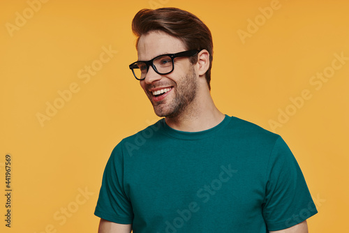 Cheerful young man in eyeglasses looking away and smiling while standing against yellow background © gstockstudio