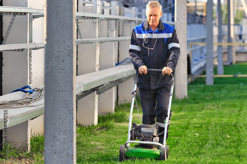 Fotografie A pensioner in working clothes works with a lawn mower on the territory of the production base on a sunny day