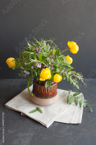 Spring bouquet. Yellow flowers of kupava and lilac flowers and green carved leaves of mouse peas in a clay vase on a gray background. photo