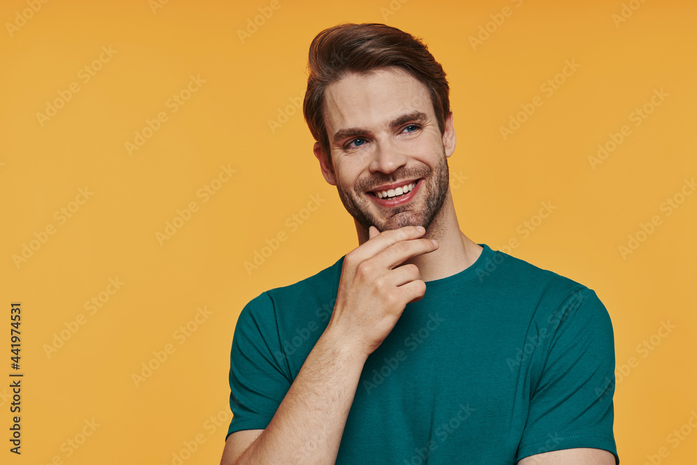 Thoughtful young man holding hand on chin and smiling while standing against yellow background Thoughtful young man holding hand on chin and smiling while standing against yellow background