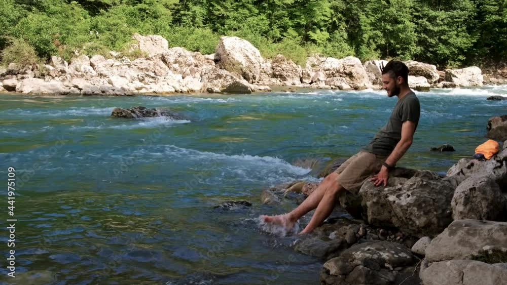 Tourist dangles his bare legs in cold water and laughs. Handsome Caucasian man with beard and dreadlocks is sitting on large stone and washing his feet in cool, stormy mountain river of bluish hue. Tourist dangles his bare legs in cold water and laughs. Handsome Caucasian man with beard and dreadlocks is sitting on large stone and washing his feet in cool, stormy mountain river of bluish hue.