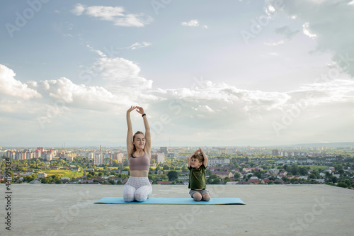 Mother and son doing exercise on the balcony in the background of a city during sunrise or sunset, concept of a healthy lifestyle © volody10