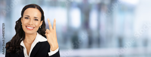 Businesswoman in black confident suit showing two fingers, second gesture, over blurred modern office interior background. Happy smiling gesturing brunette woman, indoors. Business success concept. © vgstudio