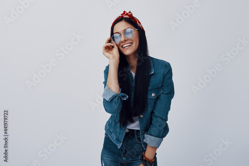 Attractive young woman in bandana adjusting eyewear and smiling while standing against grey background
