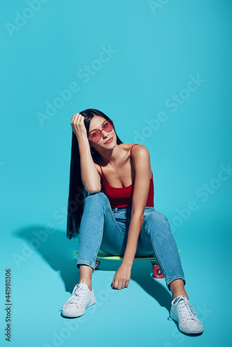 Attractive young woman looking at camera while sitting on skateboard against blue background
