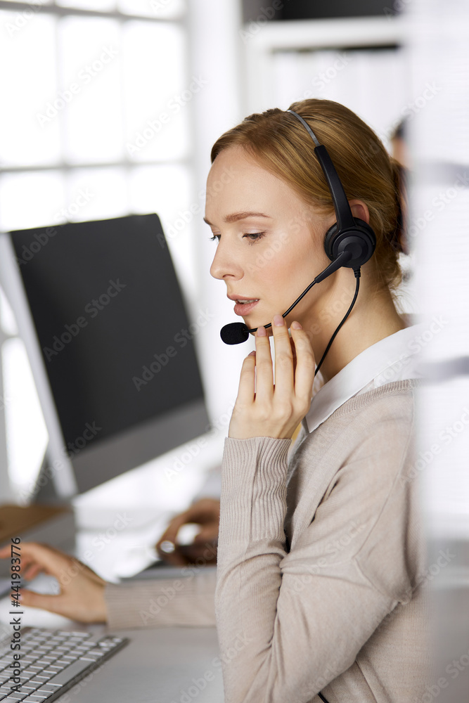 Friendly businesswoman talking by headset while sitting in office. Call center and diverse people group in business Friendly businesswoman talking by headset while sitting in office. Call center and diverse people group in business