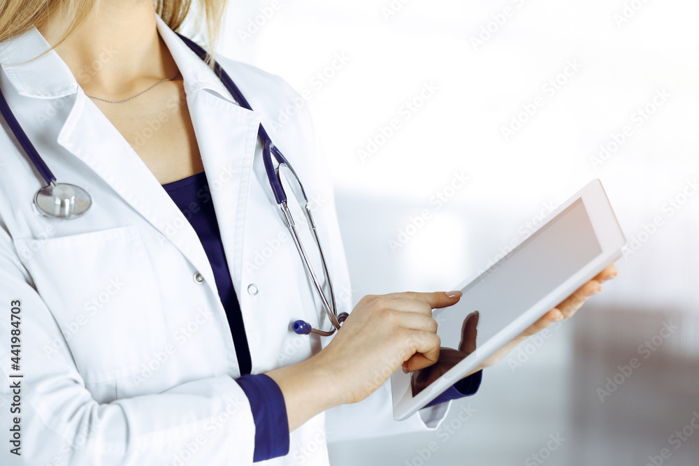 Unknown woman-doctor is holding a tablet computer in her hands, while standing in a sunny clinic cabinet. Female physician at work, close-up. Perfect medical service in a hospital. Medicine concept Unknown woman-doctor is holding a tablet computer in her hands, while standing in a sunny clinic cabinet. Female physician at work, close-up. Perfect medical service in a hospital. Medicine concept