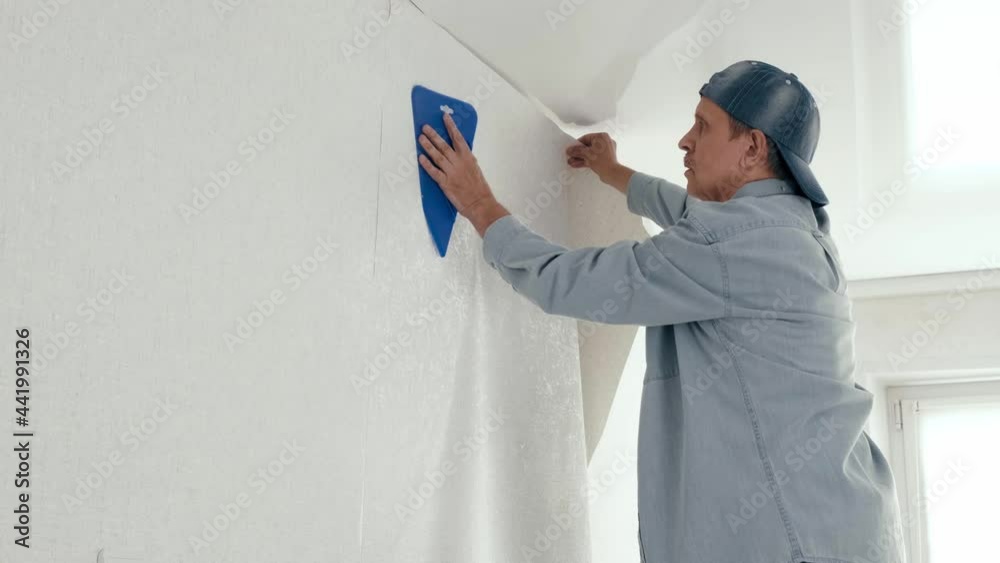 4K. Man with spatula smoothing wallpaper on the wall, smoothes fresh pasted wallpaper. Renovation of the apartment. interior design and Room renovations at house. to paste Wallpaper on the wall