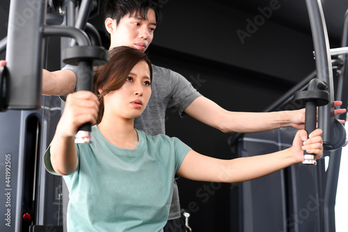  マシンで筋トレ（チェストプレス）をするアジア人女性と補助をする男性トレーナー