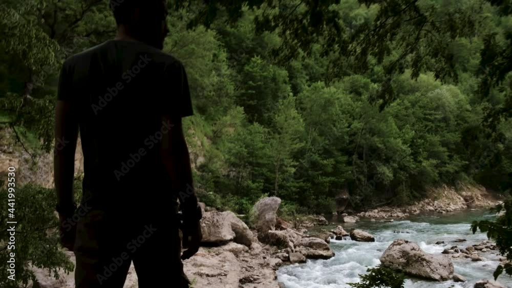 Caucasian man with dreadlocks stands on cliff on observation deck and looks at beautiful stormy cold mountain river flowing through stone gorge below. Explore the world and enjoy life. Caucasian man with dreadlocks stands on cliff on observation deck and looks at beautiful stormy cold mountain river flowing through stone gorge below. Explore the world and enjoy life.