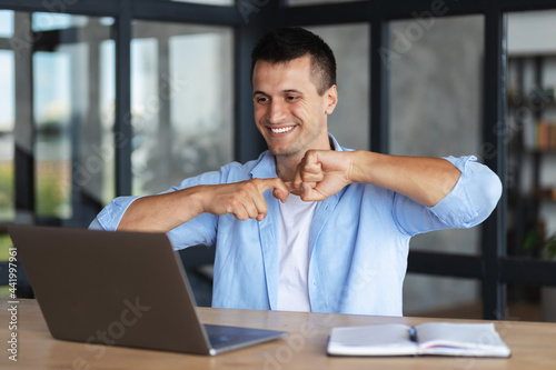 Deaf or hard hearing happy smiling young caucasian man uses sign language while video call using laptop while sitting at a desk in a home office