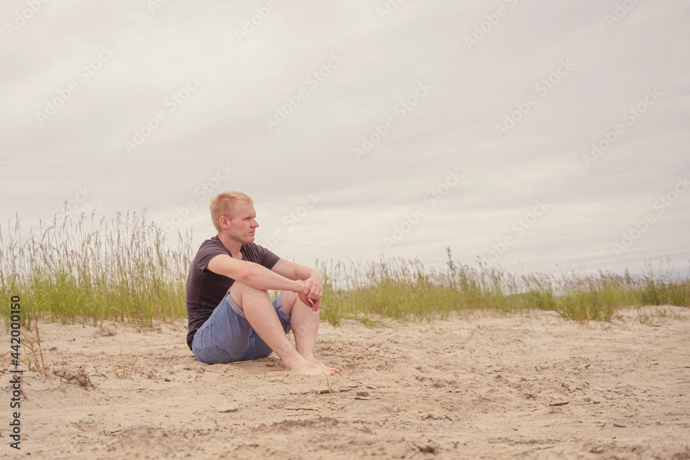 A young Caucasian man in a gray T-shirt and blue denim shorts is sitting on the sand by the river and looking into the distance. The concept of outdoor recreation, relaxation.