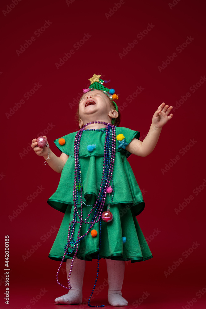 little girl in a Christmas tree costume stands on a red background. The child is very upset, crying and screaming, very unhappy.