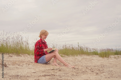 A young Caucasian girl in a red shirt and blue denim shorts sits on the sand by the river and looks into an open book. The concept of outdoor recreation, relaxation. © Ольга Холявина