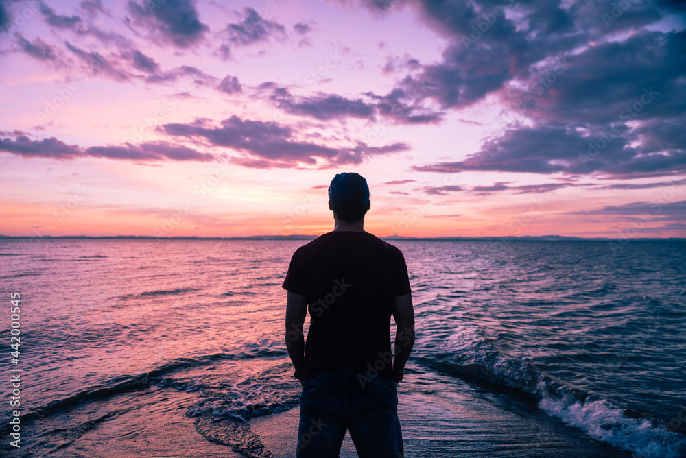 Young traveler man with his back turned at sunset, with a colourful background of the sea and a beautiful sky.