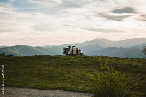 Back view of a Spanish man and a dog admiring nature while sitting on a camper van parked on a hill