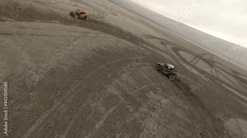 Large buggy automobiles drift on dried salt lake territory with brown sand leaving circle traces against hill silhouettes. Aerial top view on freestyle fpv drone photo