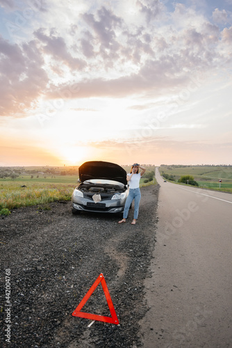 A frustrated young girl stands near a broken-down car in the middle of the highway during sunset. Breakdown and repair of the car. Waiting for help.