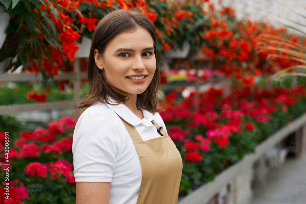 Close up portrait of smiling attractive young woman in special beige uniform standing in greenhouse on flowers background. Concept of work and care for flowerpots. Close up portrait of smiling attractive young woman in special beige uniform standing in greenhouse on flowers background. Concept of work and care for flowerpots.