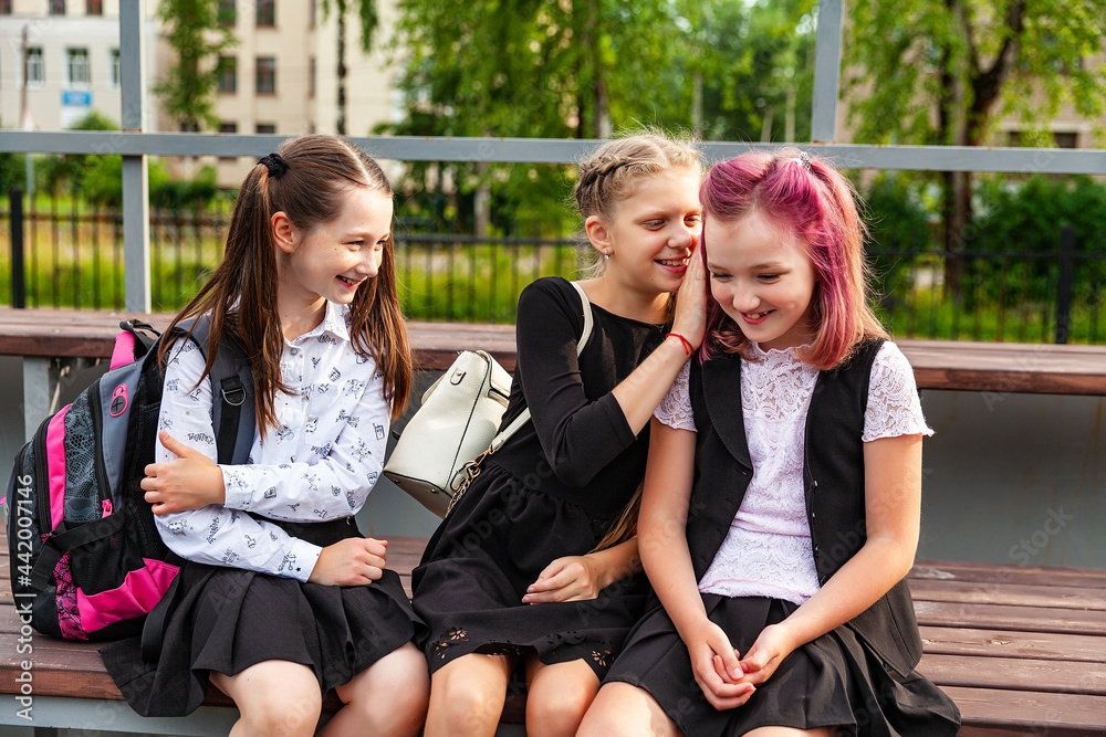 Three schoolgirls have fun chatting, sitting on a bench in the school playground outdoors. School black and white clothing, education, break in lessons Three schoolgirls have fun chatting, sitting on a bench in the school playground outdoors. School black and white clothing, education, break in lessons